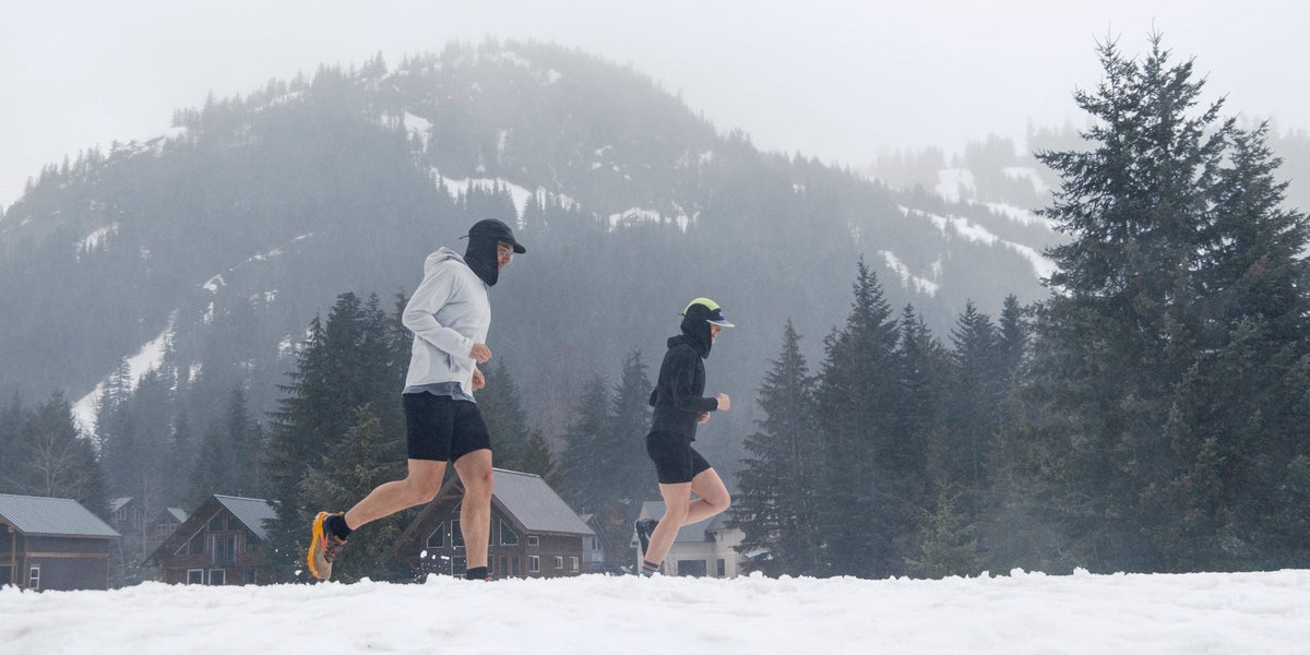 Two runners in snow beside mountain cabins in Fractel winter running gear, our winter running headwear range