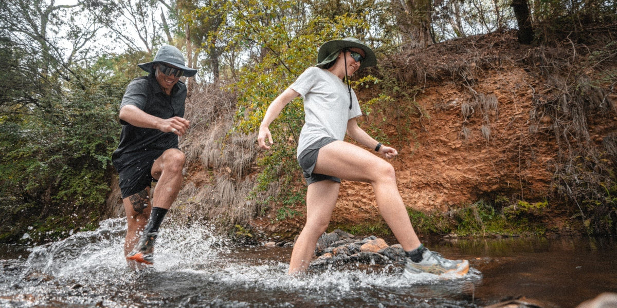 Two trail runners crossing a creek in Fractel adventure hats, an adventure hat with neck cape built for the trail