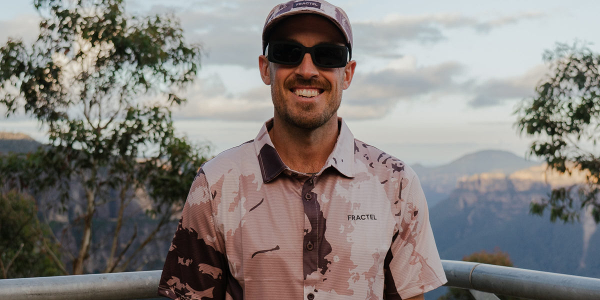 Smiling runner in a Fractel cap and earth-toned printed collared adventure shirt at a mountain lookout