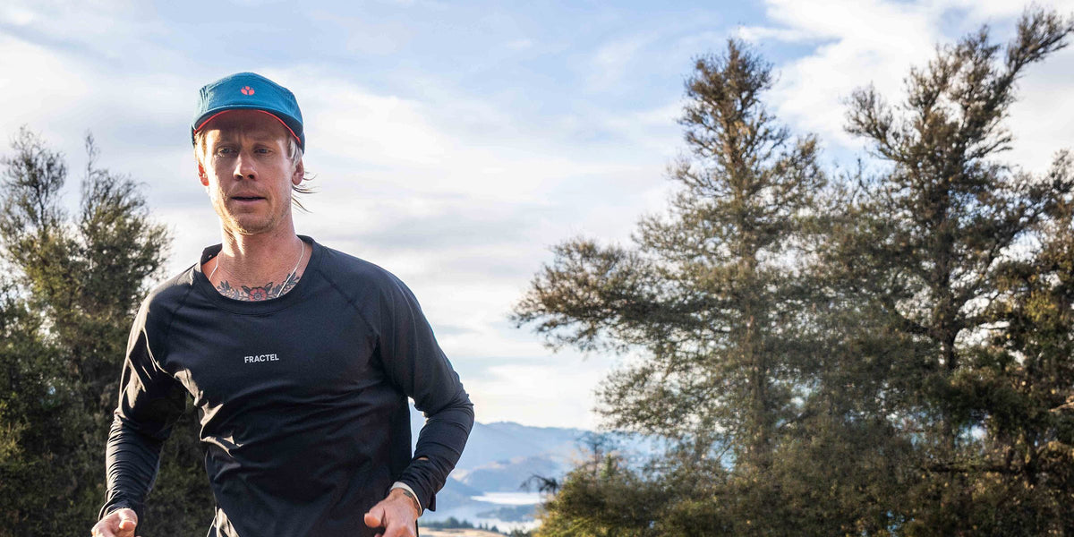 Runner in a black Fractel long sleeve running shirt and teal cap on a ridge with distant mountains