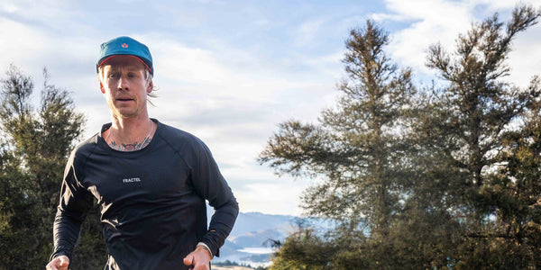 Runner in a black Fractel long sleeve running shirt and teal cap on a ridge with distant mountains