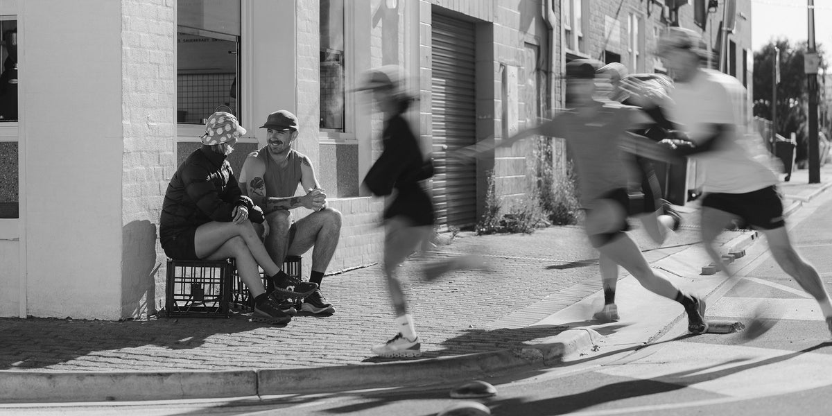 Black and white photo of runners in motion past two friends seated on milk crates, shop running gear online at Fractel