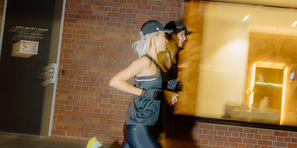 Two women in grey Fractel womens running singlets and caps running past a brick wall at night