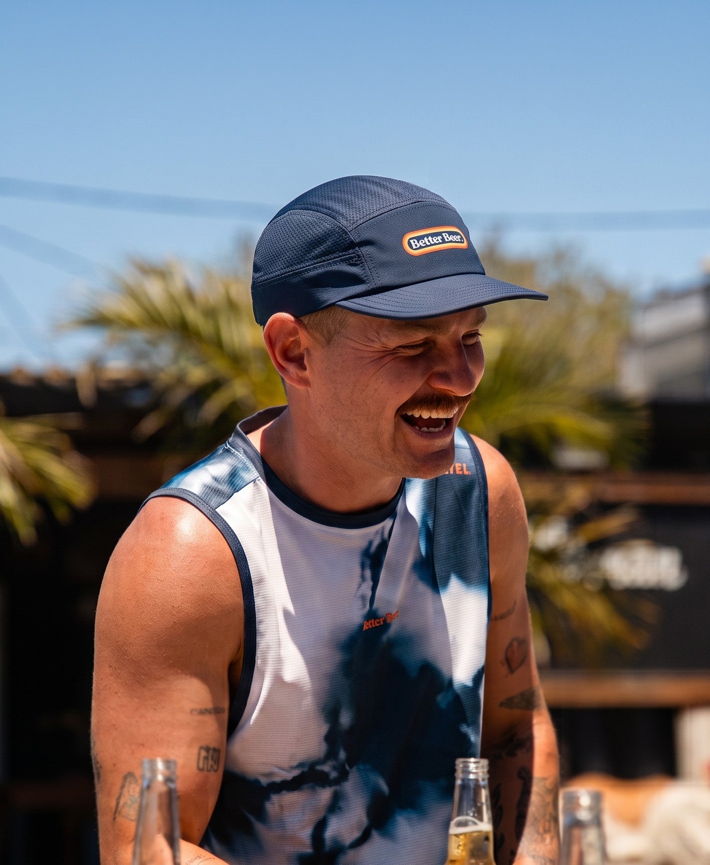 A man outdoors wearing a navy blue cap with a visible 'Better Beer' logo and a tie-dye singlet, smiling while holding beer bottles in a sunny outdoor setting.